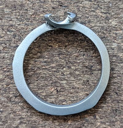 The freshly 3D-printed titanium ring on a wooden surface, matte grey with a rough, unfinished claw at the top